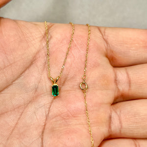 14K yellow gold solitaire pendant featuring a 5x3 mm emerald-cut lab emerald, showing prong setting and fine Singapore chain texture. 