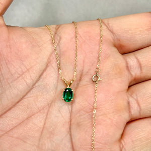 Oval medium-dark green lab created emerald pendant in 14K yellow gold, showing smooth prong setting and fine-scale solitaire proportions on a delicate chain. 