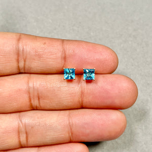 Pair of princess-cut Swiss blue topaz stud earrings in polished 14K yellow gold, showing medium blue stones held by four prongs with clean geometric edges and compact 5 mm scale. 