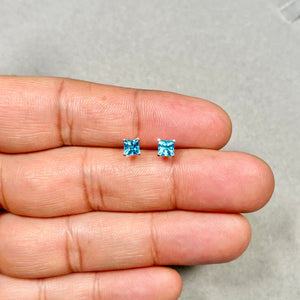 Pair of medium blue princess-cut Swiss Topaz stud earrings in 14K white gold, 4 mm stones, rhodium finish, displaying geometric sparkle. 