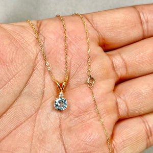 14K yellow gold aquamarine pendant featuring a 6 mm light blue round stone with single diamond accent, open prong setting, shown on fine Singapore chain for scale. 