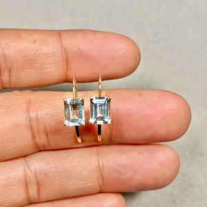Pair of emerald-cut light blue aquamarine stud earrings in 14K yellow gold, showing prong setting detail and lever back scale against neutral background. 
