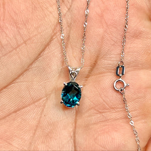 Platinum scroll pendant holding a 9x7 mm oval London Blue Topaz, dark blue gemstone secured by prongs on a white gold chain. 