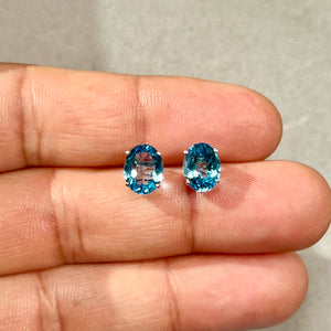 Oval Swiss Blue Topaz stud earrings in 14K white gold showing medium blue colour, four prong setting, smooth polished finish, and 9x7 mm stone scale on a neutral background. 