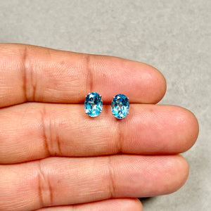 Pair of oval Swiss Blue Topaz stud earrings showing medium blue gemstones in four-prong 14K white gold settings, smooth polished metal, 8x6 mm stone size visible against neutral background. 