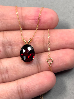 Oval red garnet solitaire pendant featuring a 12x10 mm AAA quality natural garnet in a four prong setting, crafted in solid 14K yellow gold with an elegant 18 inch Singapore chain and secure spring ring clasp. 