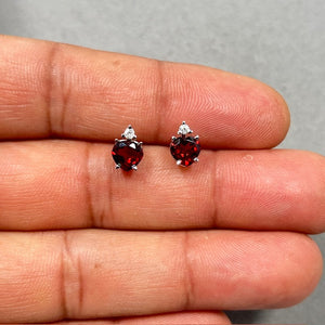 Heart-shaped natural garnet and diamond stud earrings crafted in 14K white gold with rhodium finish, featuring 1.66 carat total garnet weight and 0.06 carat round diamond accents. Elegant red gemstone studs with prong setting and secure push-back closure, designed for romantic occasions and everyday luxury wear. 