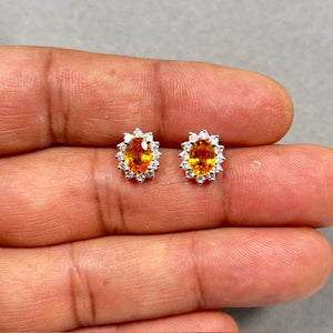 Oval yellow sapphire and diamond cluster earrings crafted in 14K white gold with rhodium finish, featuring natural 7x5 mm heated and beryllium treated sapphires totaling 1.76 carats, surrounded by 24 round cut SI-H diamonds totaling 0.48 carats in secure prong setting, elegant halo stud design with push backs, luxury fine jewelry earrings for women with vibrant golden hue and brilliant sparkle. 