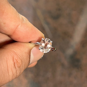AAA oval Morganite solitaire ring in 14K white gold featuring a 10x8mm faint pink gemstone weighing approximately 2.00–2.50 carats. The prong-set center stone is enhanced by an elegant scroll-style band with rhodium finish, offering a romantic blush tone and refined craftsmanship in size 7.5. 