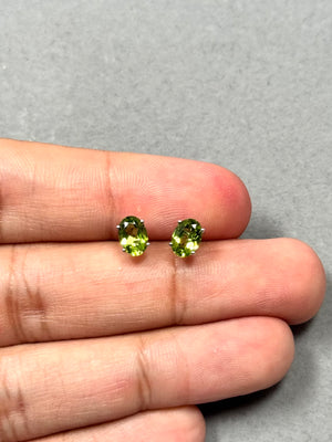 Oval peridot stud earrings in solid 14K white gold with rhodium finish, featuring natural AAA peridot gemstones (7x5 mm) in a classic four-prong setting. These 1.55–1.97 Ct green gemstone studs offer bright clarity and elegant sparkle with secure push-back closures, perfect for everyday jewellery styling. 