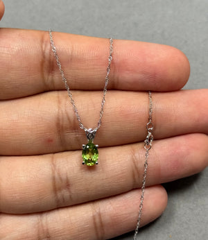Dainty peridot scroll pendant in 14K white gold featuring a natural AAA oval 7x5mm gemstone in a prong setting. Finished with rhodium for lasting shine, this elegant women’s necklace includes an 18-inch Singapore chain. A minimal green gemstone design perfect for everyday wear and refined styling. 